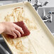 A hand using a red sponge to wash a Baker's Mark ivory aluminum sheet pan over a sink.