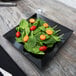 A black GET Siciliano square plate with spinach and tomatoes on a table.