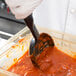 A person in a white coat using a Vollrath High Heat Perforated Oval Nylon Spoodle to scoop red sauce into a container.