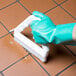 A hand in a green glove using a Carlisle white grout brush to clean tile.