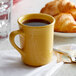 A yellow Tuxton Concentrix mug filled with a brown drink on a white napkin next to croissants.