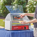 A man using an Avantco hot dog roller grill to cook hot dogs on a table with a blue tablecloth and a red sign.