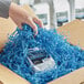 A hand holding a package of Spring-Fill Sky Blue Crinkle Cut paper shred in a cardboard box.