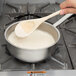 A person using a Vollrath stainless steel saucier pan to stir white liquid with a wooden spoon.