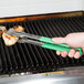 A person holding a pair of Vollrath VersaGrip tongs with green Kool Touch handles over food on a grill.