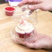 A person's hand holding a cupcake with white frosting and sprinkles in a clear plastic container with a hinged lid.