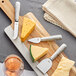 A piece of cheese on a wooden cutting board with Acopa stainless steel cheese knives and a glass of wine.