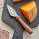 An Acopa cheese knife cutting a block of cheese on a cutting board.