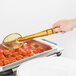A person holding a Cambro amber high heat solid salad bar spoon over a tray of food.