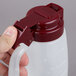 A hand holding a Tablecraft plastic dispenser jar with a maroon lid.