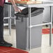 A person standing next to a Lavex Slim Gray rectangular under-counter trash can.