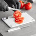 A person using a white Choice cutting board to cut a tomato with a knife.