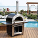 A black and silver Pinnacolo hybrid wood/gas-fired outdoor pizza oven on a outdoor table.