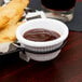 A white fluted melamine ramekin filled with brown sauce on a table with fries.