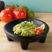 A charcoal grande molcajete filled with guacamole and tomatoes on a table.