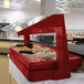 A red Cambro table top buffet with a free standing sneeze guard over food.