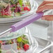 A hand holding a Baker's Mark purple silicone handle over a container of salad.