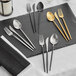 Acopa Odin brushed stainless steel flatware set with gold accents on a black table.