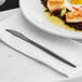 A white plate with a black Acopa Odin dinner knife on a white napkin next to food.