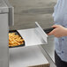 A woman using a 13 1/2" x 9 1/2" aluminum paddle peel with a black handle to remove french fries from an oven.