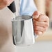 A person using an Acopa stainless steel frothing pitcher to pour milk.