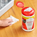 A hand using a Sani Professional white paper towel to clean a kitchen counter with a container of Sani Professional sanitizing wipes.