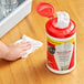 A hand using a Sani Professional sanitizing wipe to clean a red container on a table.