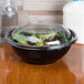 A Sabert black round bowl filled with salad on a table in a salad bar.