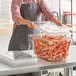 A woman in an apron using a clear Cambro food storage container to store carrots.