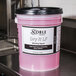 A pink bucket of Noble Chemical Dry It LF Concentrated rinse aid on a counter in a professional kitchen.