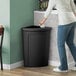 A woman throws a bag into a Lavex corner round black trash can with a black lid.