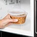 A hand holding a Pactiv translucent round deli container lid filled with food in a microwave.