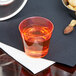 A Fineline Quenchers neon red hard plastic shot cup filled with orange liquid on a table.