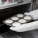 A person in gloves using a Chicago Metallic mini cake and jumbo muffin pan to bake muffins in the oven.