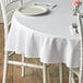 A white table with a Choice white round table cover and a white plate with a fork on it, with flowers in the background.