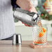 A hand using an Acopa stainless steel cocktail shaker to pour orange juice into a glass.