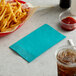 A teal 2-ply paper dinner napkin on a table with a cup of soda and a basket of french fries.