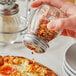 A person holding a Choice glass cheese shaker pouring pepper flakes on a pizza.