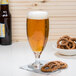 A Libbey stemmed pilsner glass filled with beer next to a bowl of pretzels.