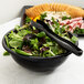 A bowl of salad in a black Fineline PET plastic bowl with black tongs.