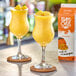 A table with two glasses of mango drinks, one garnished with orange, and a box of Island Oasis Mango Frozen Beverage Mix.