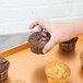 A hand holding a chocolate muffin on a Chicago Metallic copper bakery display tray.