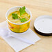 A Huhtamaki Streetside Print white double poly-paper soup cup with a vented paper lid on a table with a bowl of soup and a white spoon.