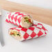 A sandwich wrapped in red and white checkered paper next to a sandwich wrapped in red and white paper.