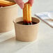 A hand dipping a Choice kraft paper take-out container into a cup of chocolate sauce with a churro.