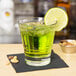 A Libbey stackable rocks glass with green liquid and a lime wedge on a black napkin.
