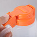 A hand holding a Tablecraft plastic dispenser jar with an orange plastic lid.
