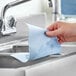A hand holding a Tork blue single fold paper towel over a sink.