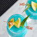 Two blue drinks on a table with Barfly copper cocktail picks topped with pineapple slices.