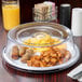 A plate of food covered with a clear Cambro plate cover on a table with a glass of orange juice.